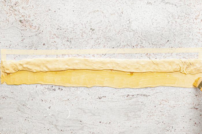 A strip of rolled-out dough is laid on a marble surface. It is being brushed with a light coating of egg wash or butter using a pastry brush. Another thin strip of dough is placed above the rolled dough.