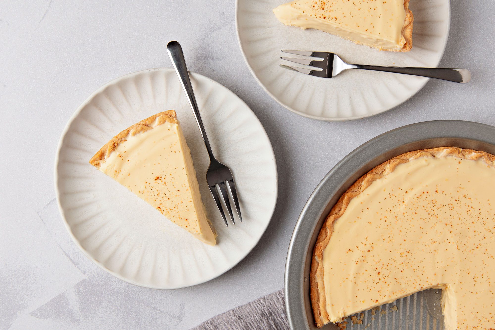 overhead shot of a pie with a light yellow custard filling and a flaky crust; a slice of pie is on a white plate with a fork beside it; another slice is on a white plate with a fork next to it; the whole pie is in a silver pie pan, with a slice missing; the plates and pan are on a white table with a grey cloth napkin in the corner of the frame