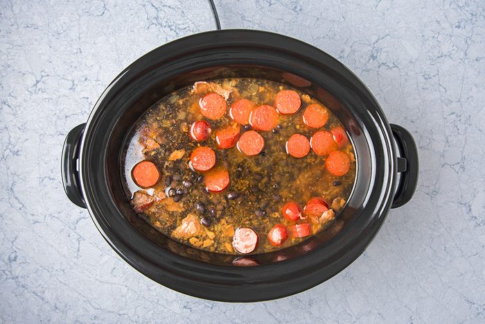 A black slow cooker filled with a stew featuring sliced sausages, tomatoes, black beans, and vegetables in a broth. The cooker is placed on a light gray marbled surface.