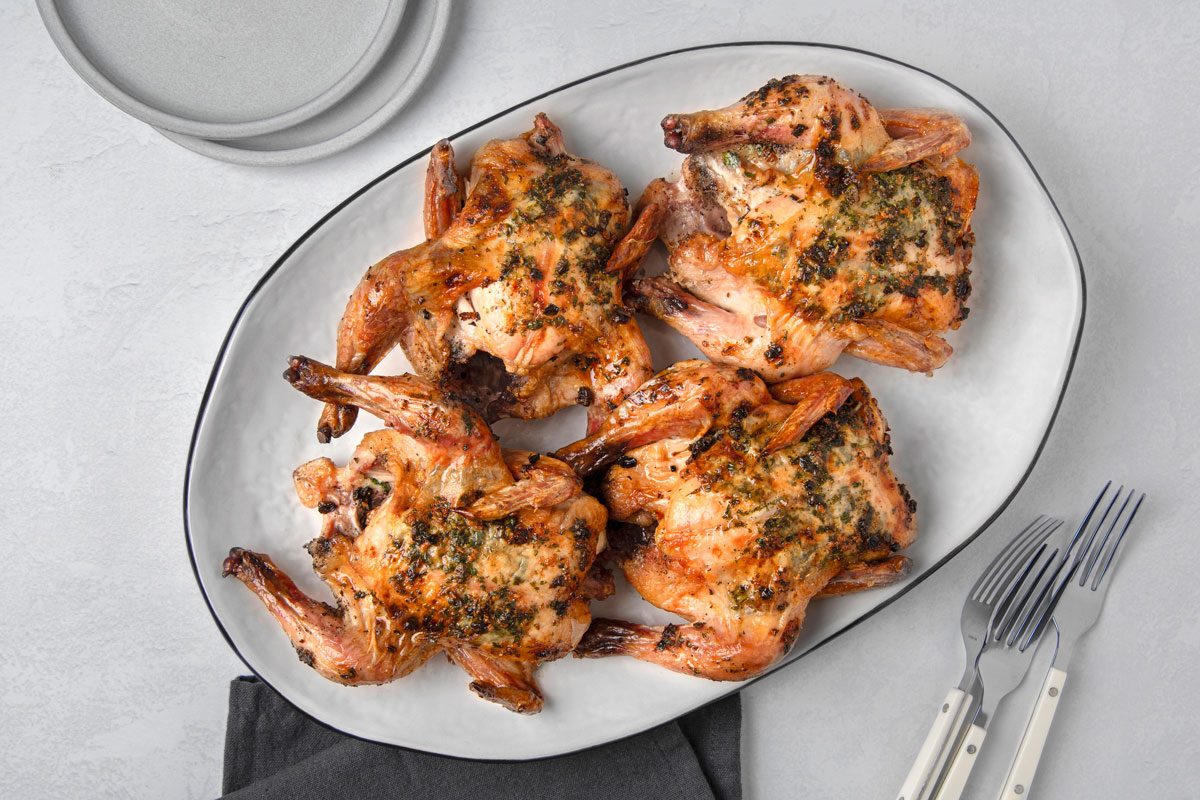 overhead shot of a platter of four roasted Cornish hens, each seasoned with herbs and spices; a grey napkin is draped over the bottom right corner of the platter, and three silver forks with white handles are arranged on the right side of the platter; the forks are partially visible and lie on the white surface beside the platter