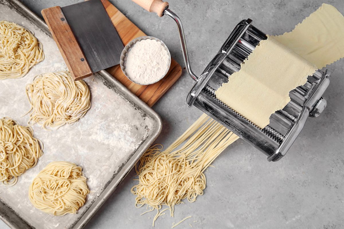 Pasta-making scene with a manual pasta machine rolling out sheets of dough and noodles onto a gray surface. Nearby, there are mounds of freshly made pasta on a floured baking sheet and a small bowl of flour on a wooden board.