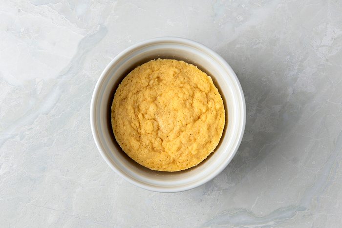 A round, fluffy cornbread muffin sits in a white ramekin on a light gray marble background.