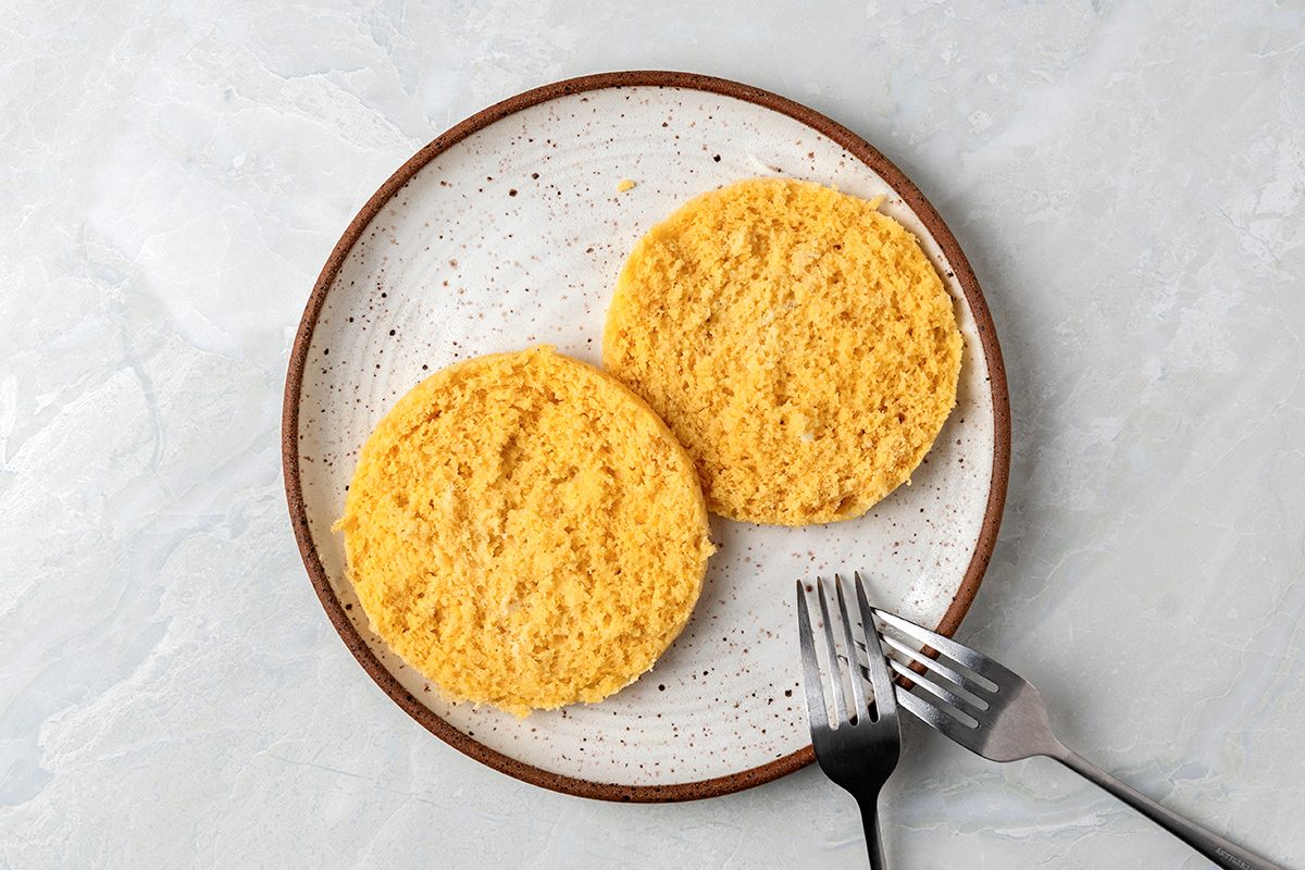 Two cornmeal arepas on a speckled ceramic plate with a silver fork and knife resting beside them on a light gray textured surface.