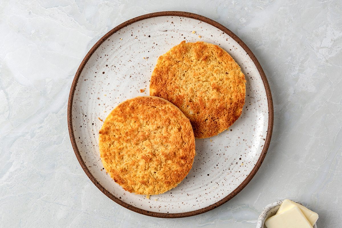 Two toasted English muffins on a speckled ceramic plate against a light gray background. A small dish with two pats of butter is partially visible in the bottom right corner.