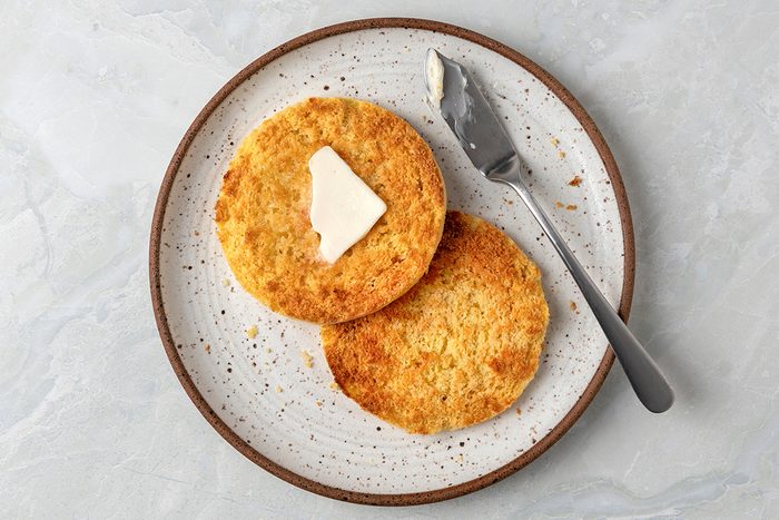 Two round pieces of toasted bread on a speckled plate, with a pat of butter melting on the top piece. A butter knife rests on the plate beside them.