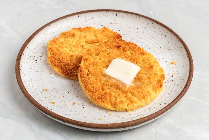 Two toasted English muffin halves on a speckled ceramic plate, with a pat of butter melting on one half. The background is a light gray surface.