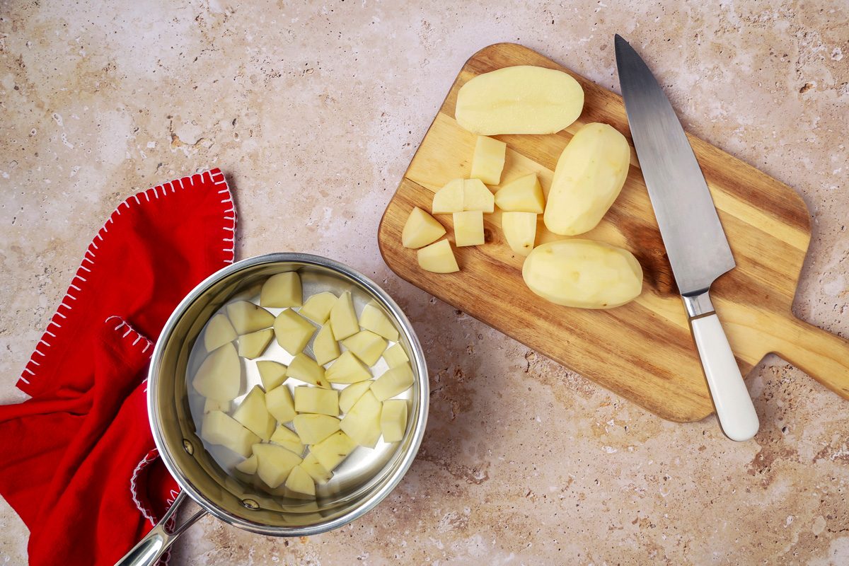 saucepan filled with water containing potatoes next to a chopping board with sliced potatoes and a knife