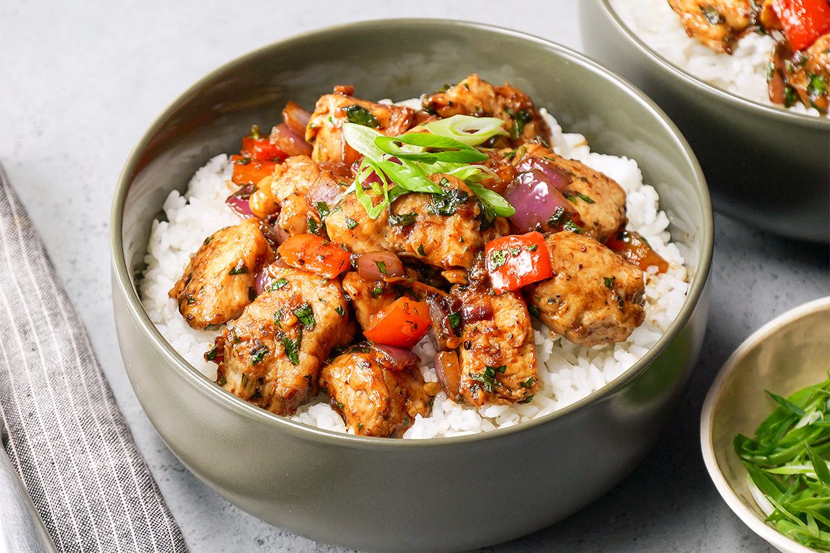 A bowl of rice topped with seasoned chicken pieces, red bell peppers, and onions, garnished with chopped green onions. The dish is served in a gray bowl on a light textured surface, accompanied by a small bowl of additional green onions.