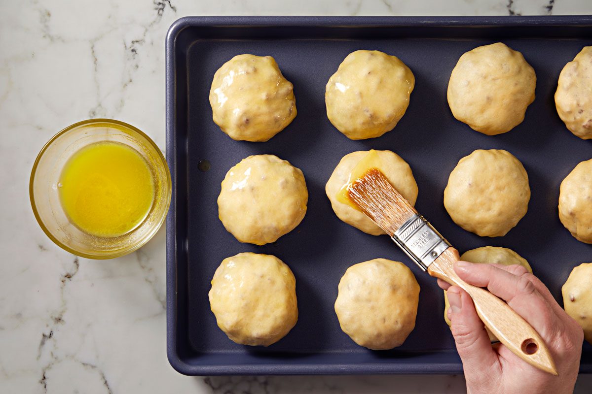 overhead shot of a baking tray filled with round dough balls; the dough is being brushed with melted butter or glaze; to the left, a small bowl containing the same liquid sits on a light marble surface