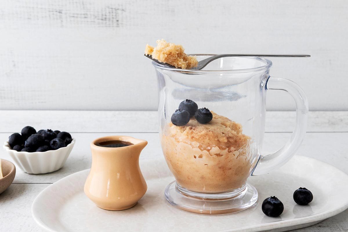 A fork holding a bite of pancake in a glass mug, topped with blueberries.