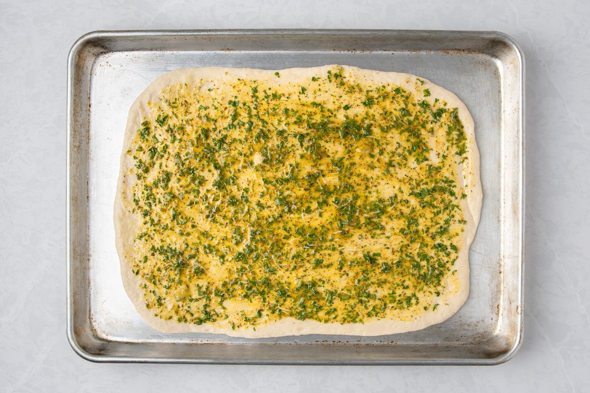 overhead shot of a rectangular piece of dough on a baking tray, the dough is covered with a yellow and green topping that resembles herbs and oil, the baking tray is metallic, a plain white surface background