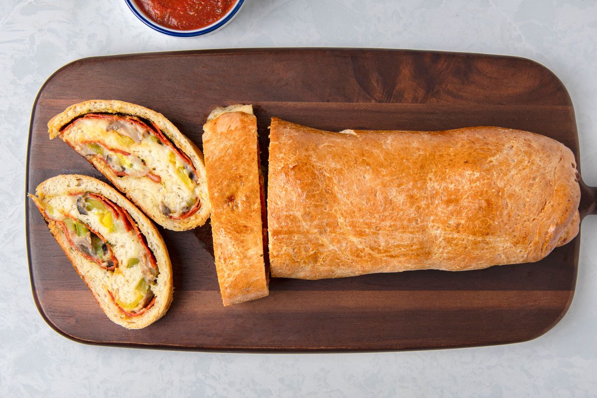 overhead shot of a sliced Pepperoni Pizza Loaf on a wooden cutting board, a small bowl containing red sauce is visible in the top left corner, the wooden board rests on a plain light surface;