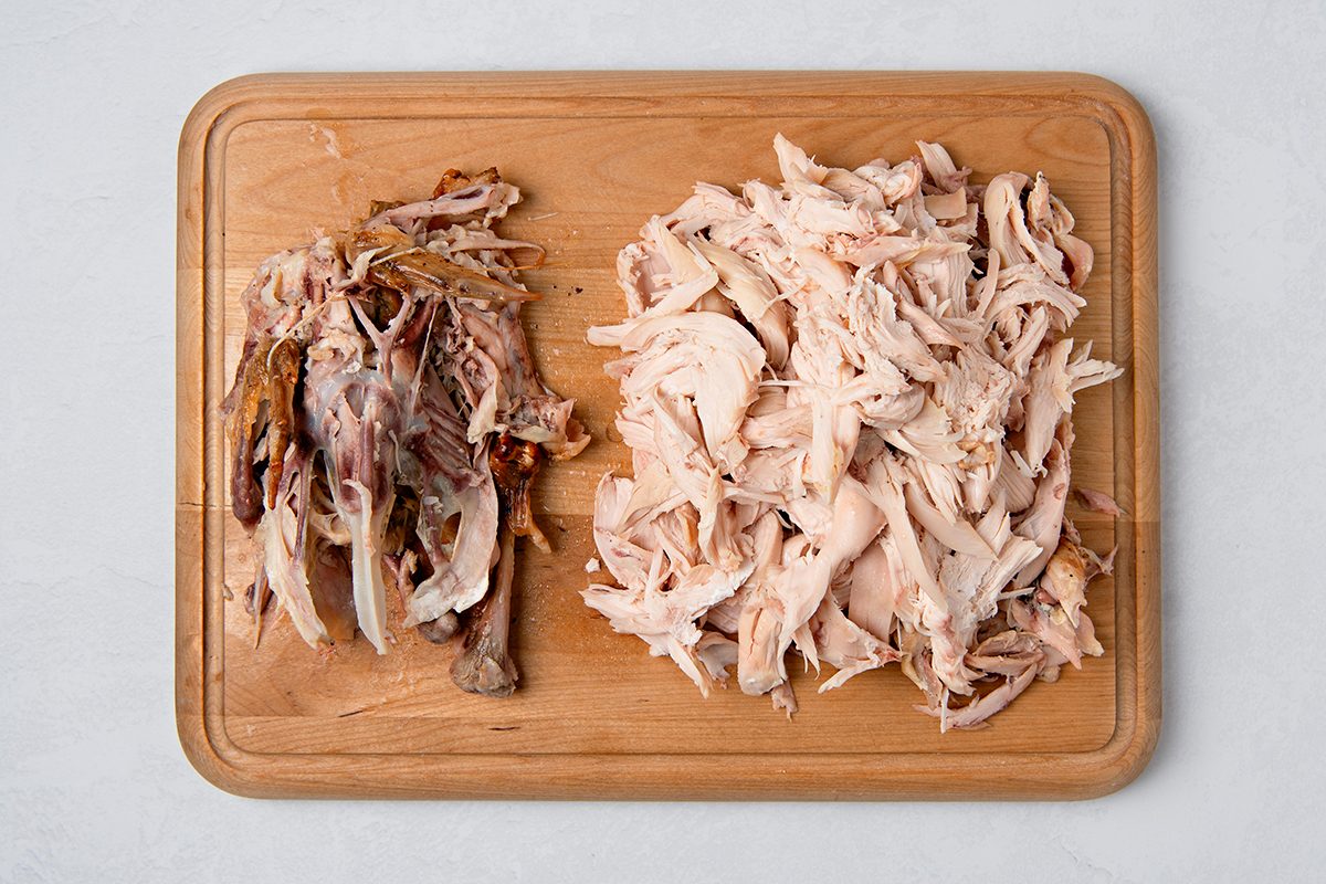 Shredded cooked chicken on a wooden cutting board, with meat on the right side and bones piled on the left. The background is a light, textured surface.
