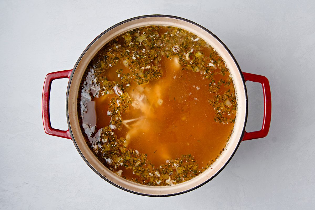 A large red pot filled with broth, visible herbs, and small pieces of vegetables or meat. The broth is a rich, light brown color. The pot is placed on a light gray surface.