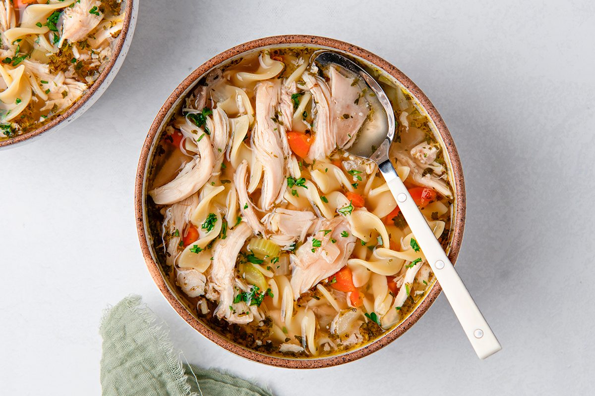 A bowl of chicken noodle soup with shredded chicken, noodles, carrots, and herbs, with a spoon resting inside. A green cloth napkin is partially visible beside the bowl on a light gray surface.
