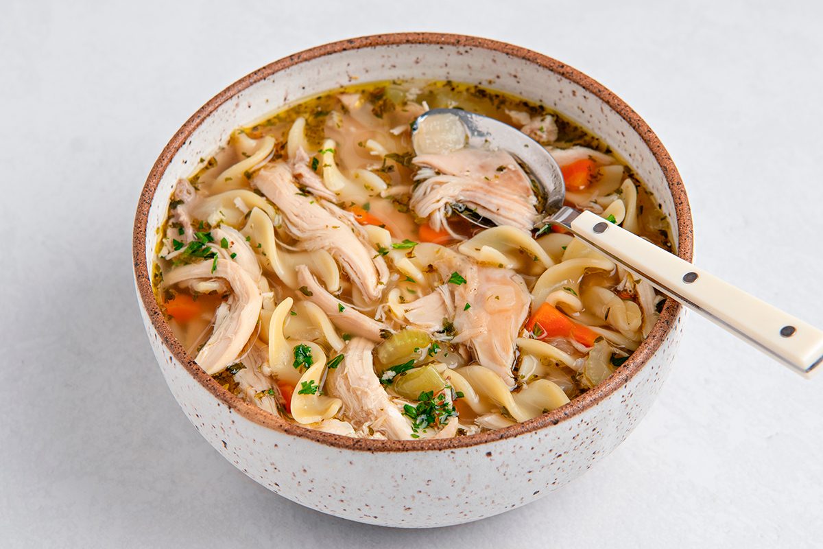 A bowl of chicken noodle soup with shredded chicken, noodles, and carrot pieces, garnished with herbs. A spoon rests in the bowl on a light background.