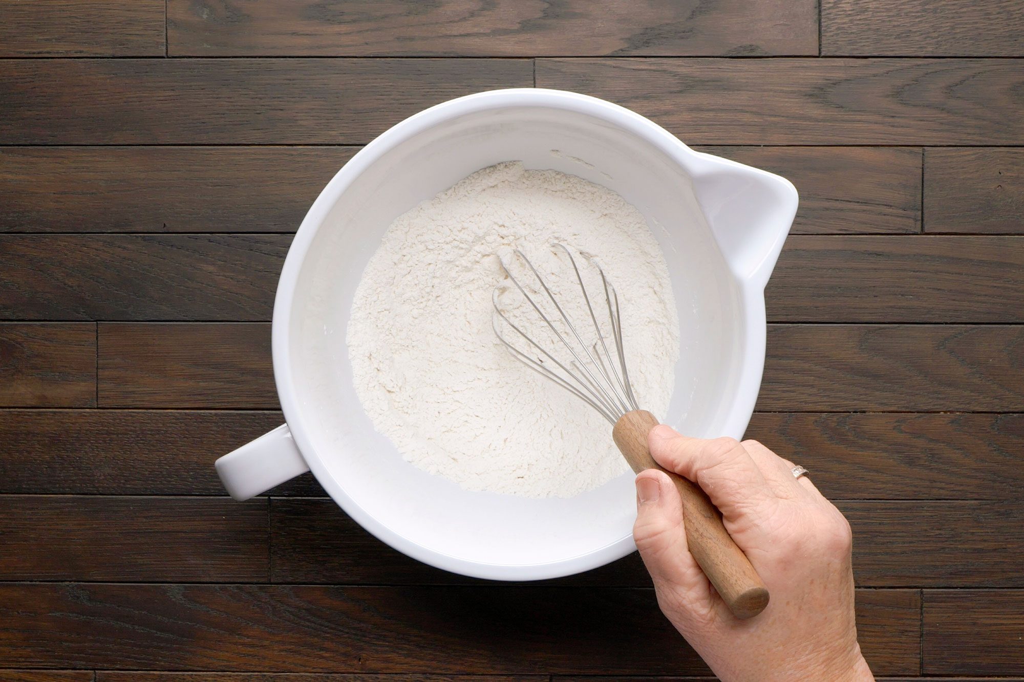 overhead shot of a bowl filled with flour, with a hand holding a whisk positioned above it; the background is wooden planks