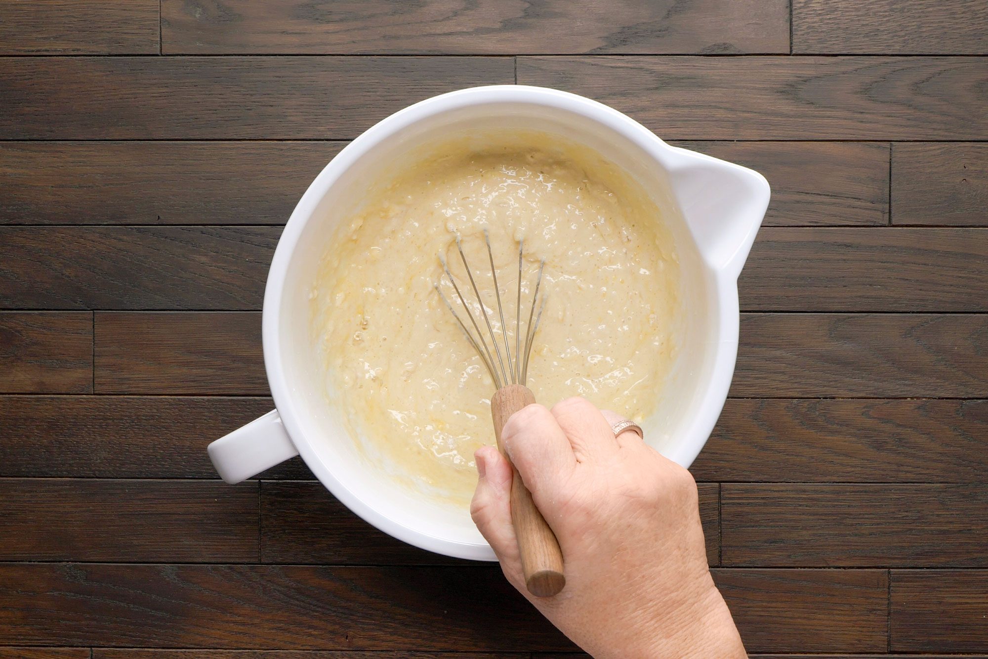 overhead shot of a mixing bowl filled with a creamy batter; stirring, a hand is visible holding a whisk with a wooden handle; the background features a dark wooden countertop