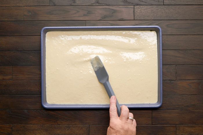 overhead shot of a baking tray filled with a creamy, light colored batter, A hand is visible holding a gray spatula, The background consists of dark wooden planks