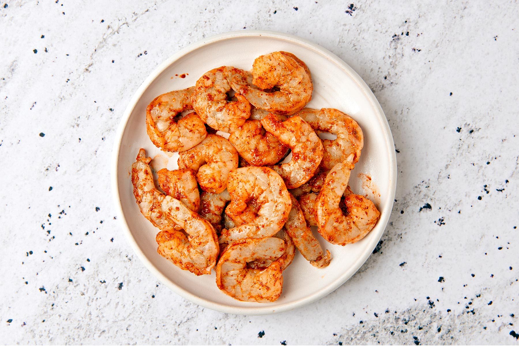 overhead shot of a white plate filled with cooked shrimp, the shrimp are seasoned with spices and are a light orange color; the plate is on a white and gray speckled countertop