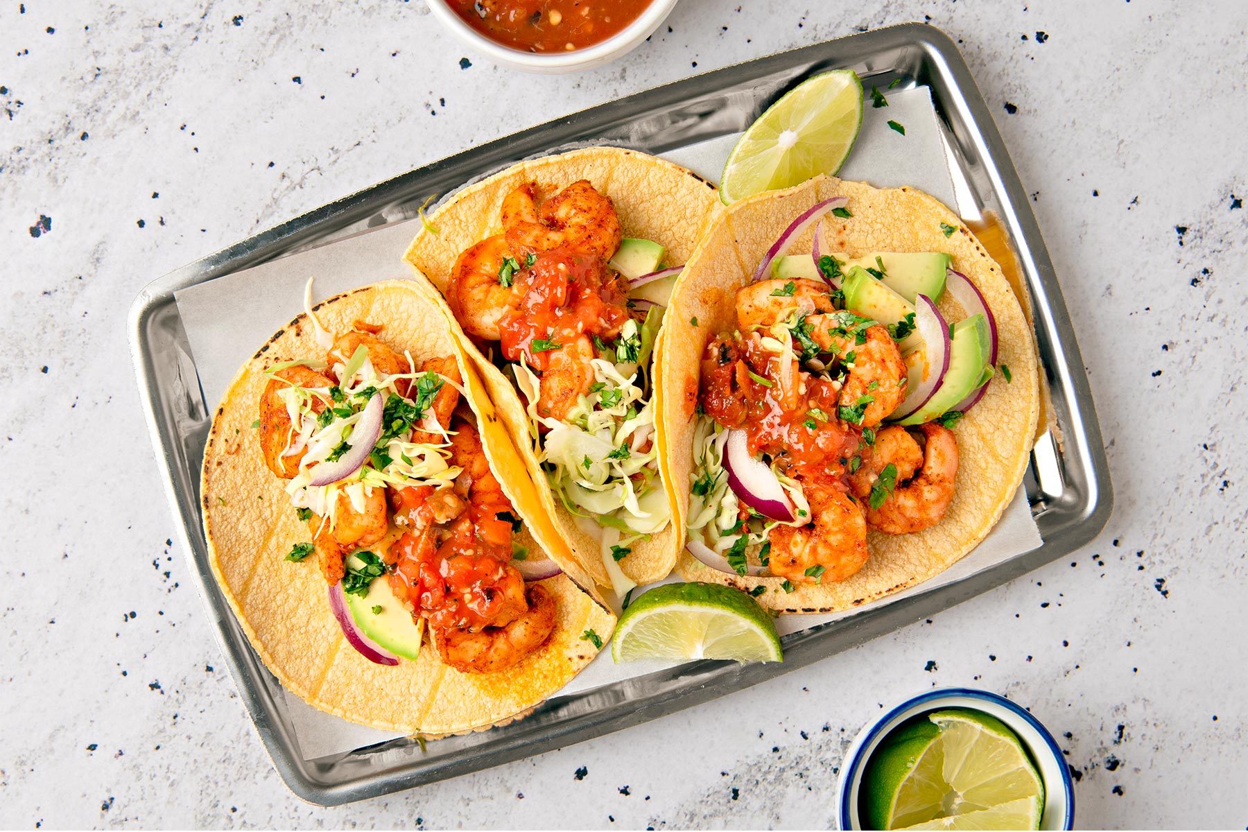 overhead shot of three shrimp tacos on a silver tray, with a white surface in the background; there is a small bowl of salsa in the top left corner of the image and a bowl of lime wedges in the bottom right corner