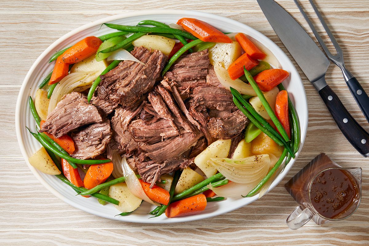 Oval plate with pot roast at the center, surrounded by cooked carrots, green beans, onions, and potatoes. A gravy-filled glass jug is beside the plate. A knife and fork rest on the wooden table.