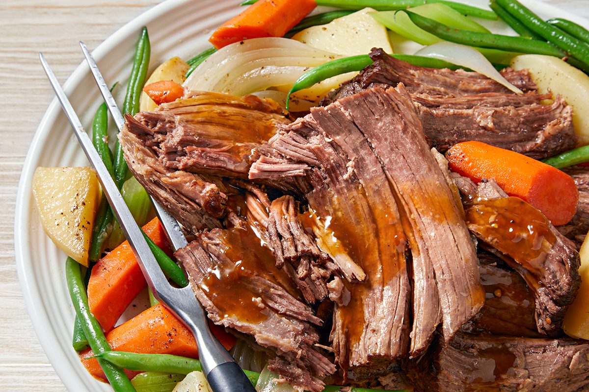 A plate of pot roast with tender meat, glazed with gravy. Surrounding the roast are carrots, green beans, sliced onions, and chunks of potato. A metal carving fork rests on the plate.