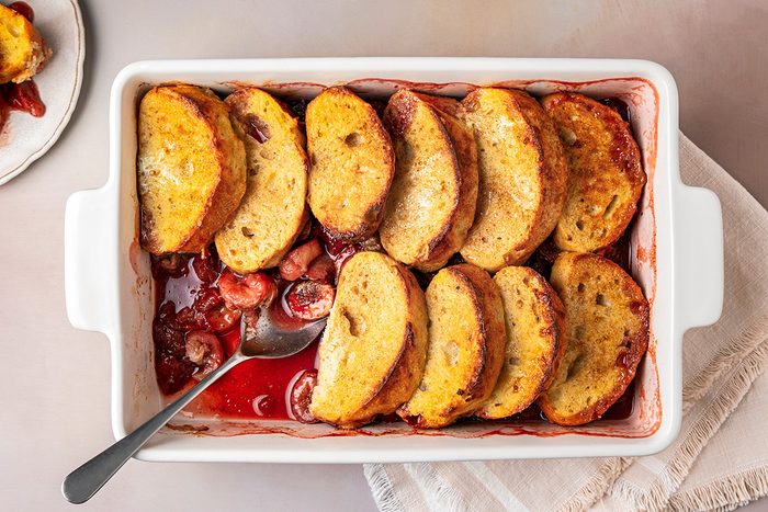 A rectangular baking dish with slices of French toast arranged on top of a layer of mixed berries. A serving spoon is placed in the dish, and a small amount of the fruit is visible at the side.