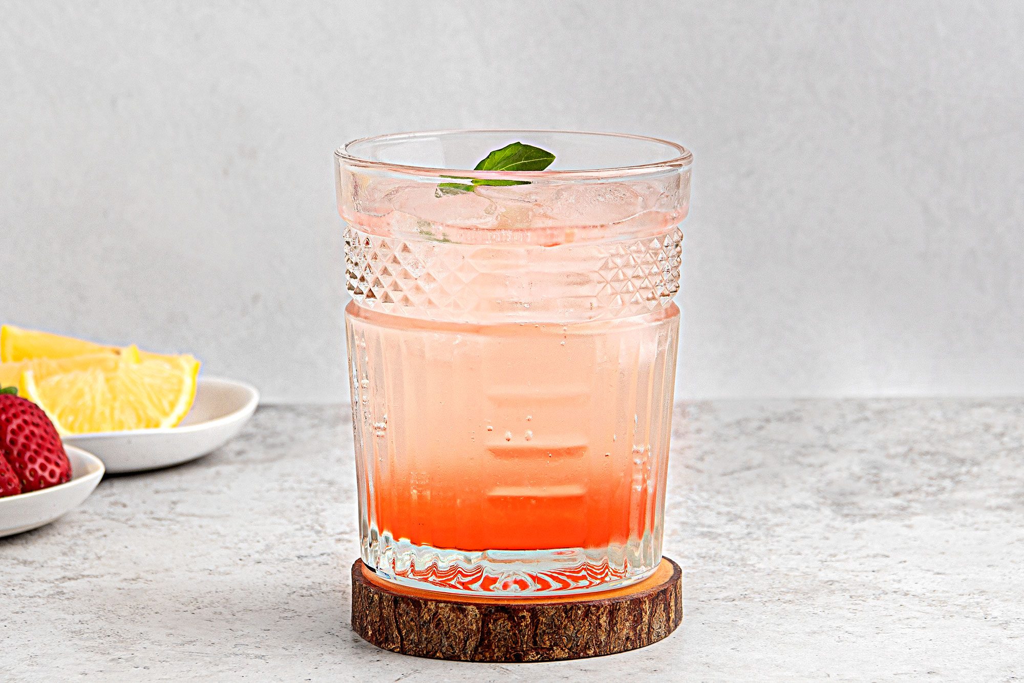 Table view shot of Strawberry-Basil Mocktail; served into rock glass filled with ice; Top with club soda and additional basil leaves; on wood coaster; strawberry on plates; lime slices on another plate; grey surface;