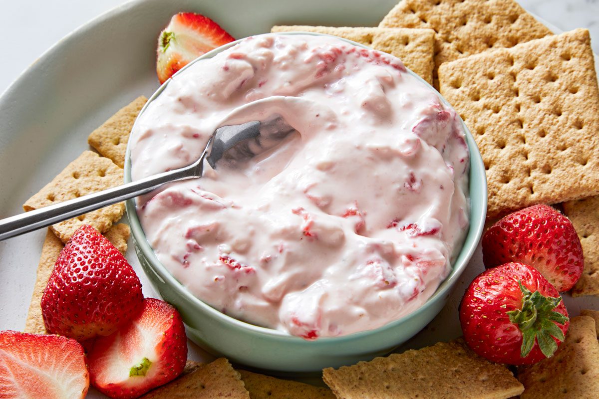 3/4th shot of a white plate filled with crackers, strawberries, and a bowl of strawberry dip; the plate is on a marble surface; there is a silver spoon in the bowl of dip