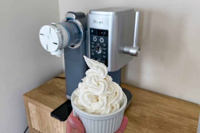 A person holds a white ceramic bowl filled with soft serve ice cream in front of a silver Ninja ice cream maker on a wooden countertop. The ice cream is swirled high above the rim of the bowl.