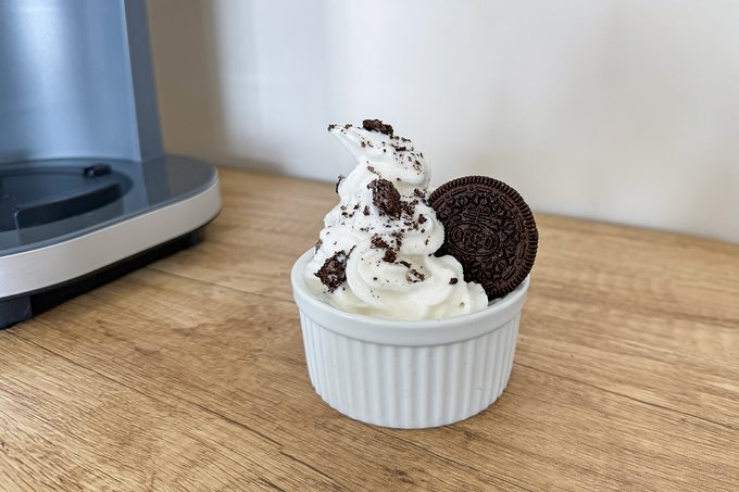 A serving of soft-serve ice cream topped with cookie crumbles and a whole chocolate sandwich cookie in a small white ceramic ramekin, placed on a wooden surface. A blurred kitchen appliance is partially visible in the background.