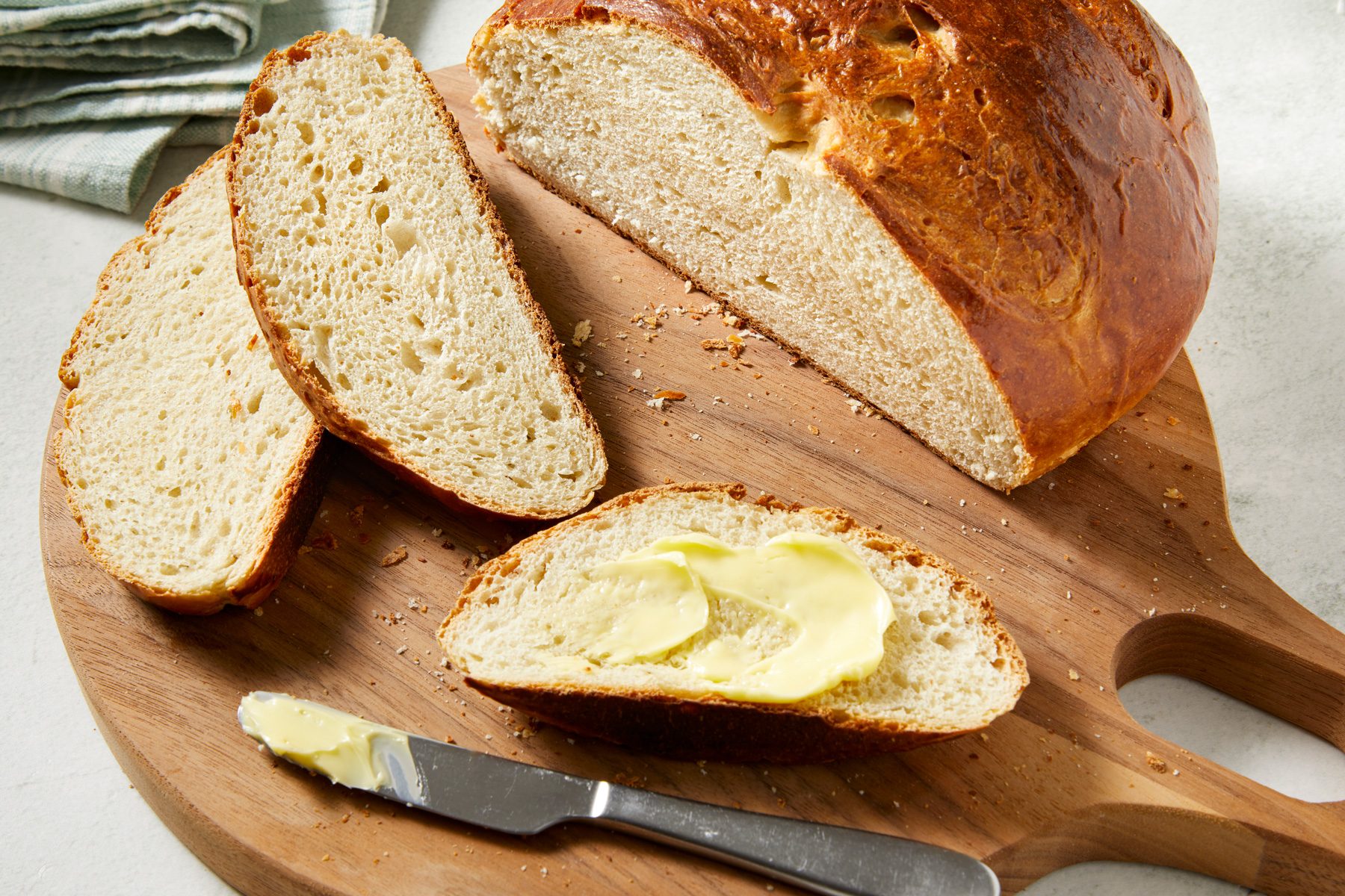 3/4th shot of baked loaf of Banana Yeast Bread, displayed on a wooden cutting board; One half of the loaf is sliced, Several slices are laid out beside the loaf, On one slice, a layer of butter is spread generously; In the background, a folded patterned kitchen towel is visible