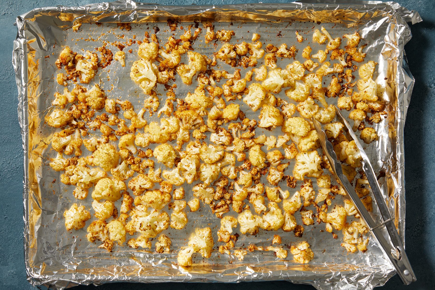 Overhead shot of Bake until cauliflower is tender and browned about 20 minutes; stirring halfway; tongs; dark blue surface;
