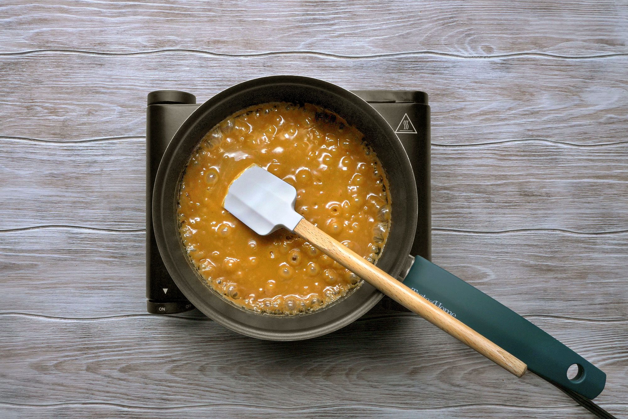 overhead shot of a pot of caramel being cooked on a portable electric stovetop, likely for making a dessert; The pot is filled with golden-brown caramel, bubbling gently as it simmers; A white silicone spatula is resting in the caramel, with a wooden handle that extends out of the pot