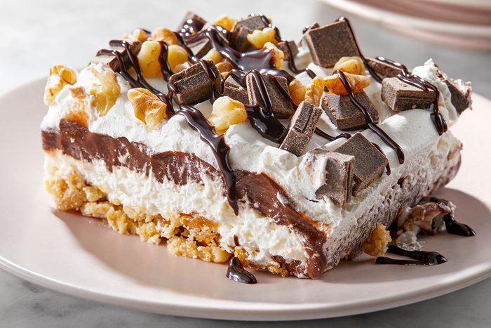 A decadent dessert bar on a plate, featuring layers of chocolate and cream filling, topped with chopped nuts, chocolate pieces, and a drizzle of chocolate syrup.