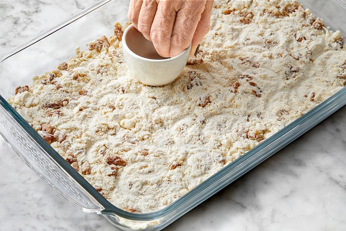 A person's hand presses a small cup into a rectangular glass baking dish filled with a crumbly mixture. The mixture includes flour and small pieces, possibly nuts, spread across a marble countertop.