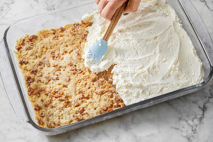 A person spreads whipped cream over a crumbly dessert topped with nuts in a glass baking dish, on a marble surface.
