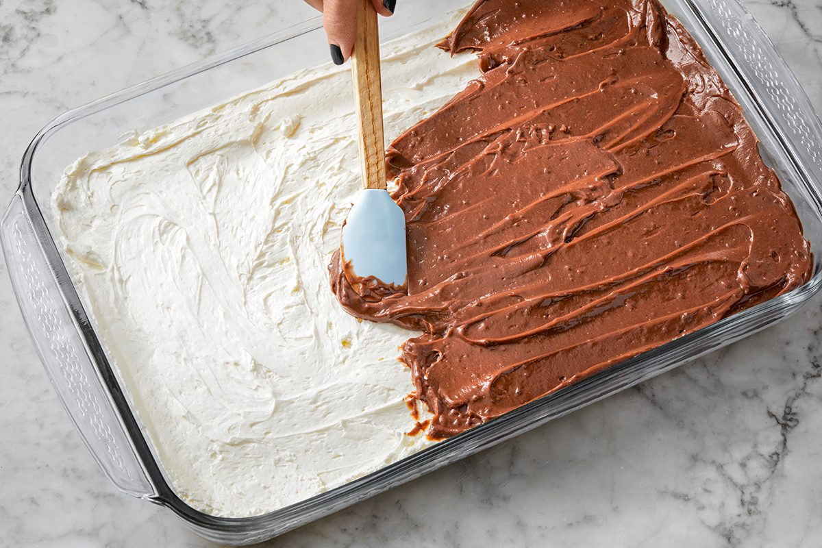 A person spreads chocolate pudding over whipped topping in a glass baking dish on a marble counter. Half of the dish is covered with white whipped topping, and the other half is being covered with the chocolate layer using a spatula.