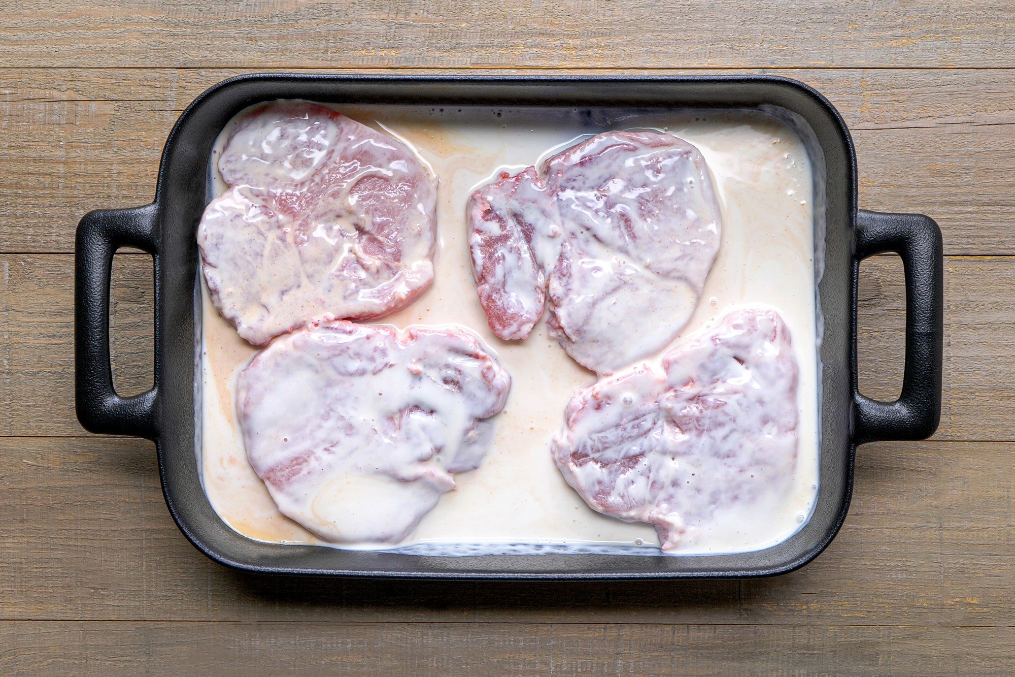 overhead shot of a rectangular baking dish containing four pieces of meat submerged in a creamy marinade, the dish is set against a backdrop of textured wooden planks