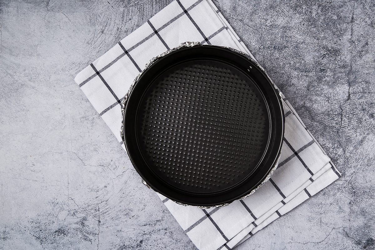 A round, black baking pan sits on a white and black checkered cloth, placed on a textured gray surface. The pan has a textured bottom and appears empty, ready for use.
