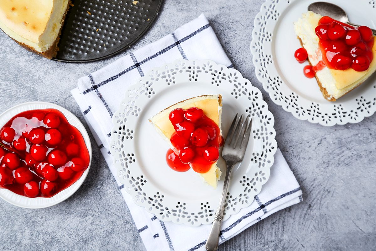 Slices of cheesecake topped with cherry sauce on ornate white plates sit on a gray surface. A white and blue checkered cloth is underneath.