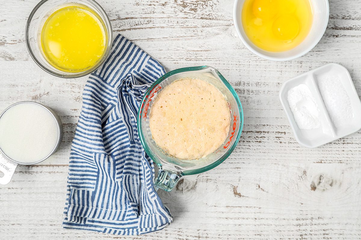 Top view of baking ingredients on a wooden surface: melted butter in a bowl, milk, eggs in a bowl, sugar, and yeast mixture in a measuring cup. A blue-striped cloth lies nearby.