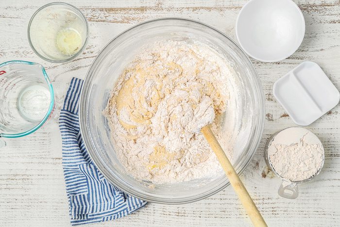 A bowl of flour and dough mixture with a wooden spoon on a white wooden table. Surrounding the bowl are a blue striped cloth, empty bowls, a measuring cup with milk, and a cup with more flour.