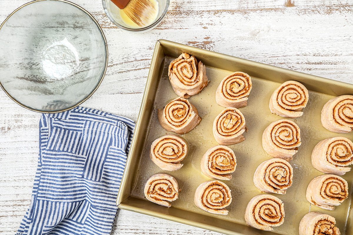 A baking pan filled with uncooked cinnamon rolls on a white wooden surface. Beside it are a clear glass bowl, a pastry brush, and a folded blue and white striped cloth.