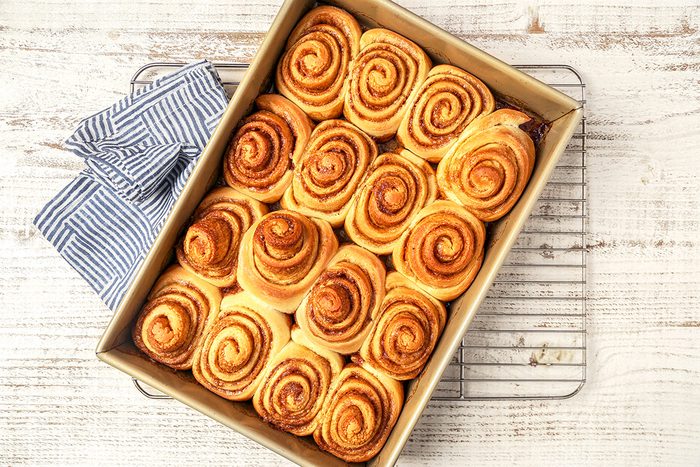 A rectangular baking dish filled with freshly baked cinnamon rolls arranged in neat rows. The rolls have a golden-brown swirl pattern on top. The dish is placed on a cooling rack, and a striped cloth is seen to the side on a light wooden surface.