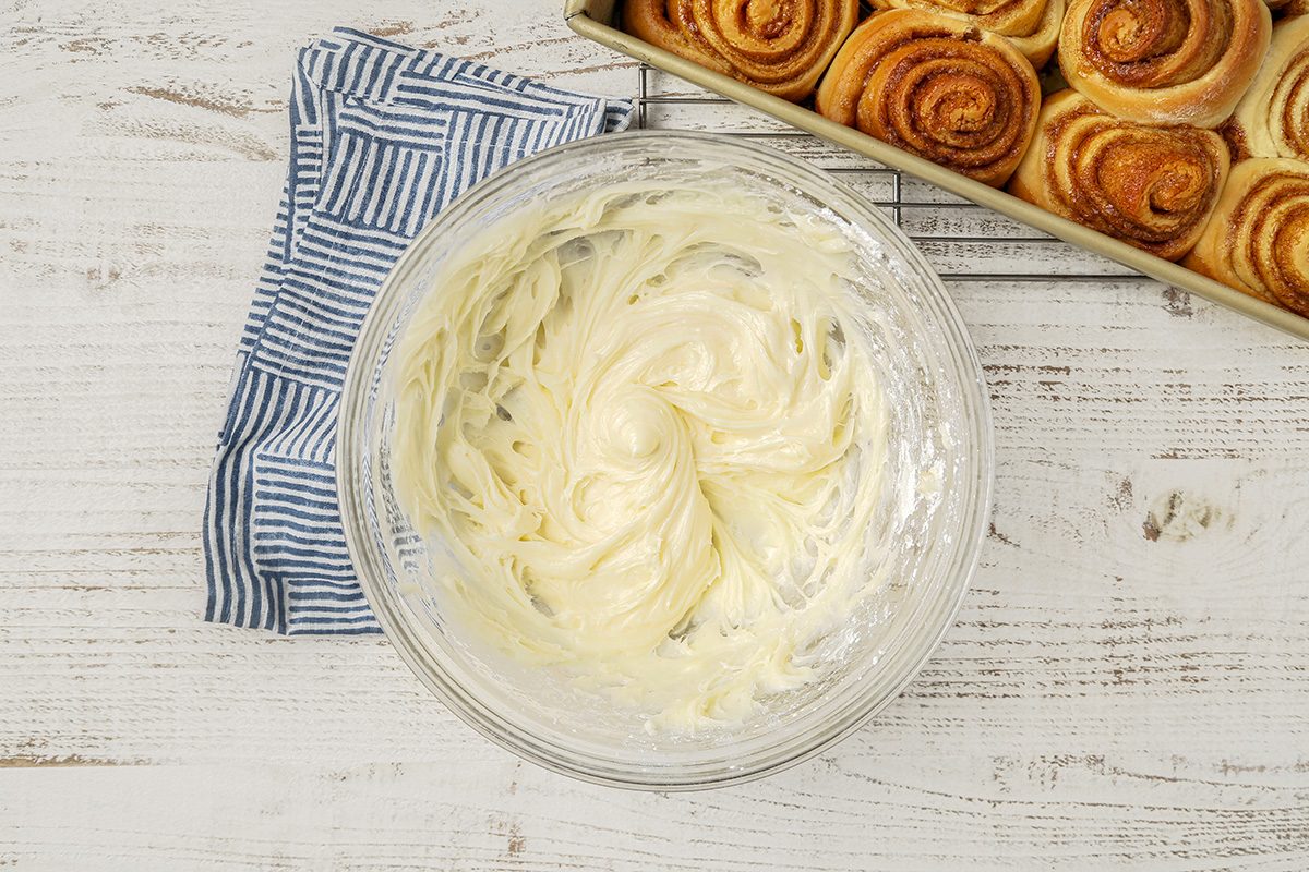 A bowl of cream cheese frosting sits on a white wooden surface next to a blue-striped cloth. To the right, a baking tray holds fresh cinnamon rolls.