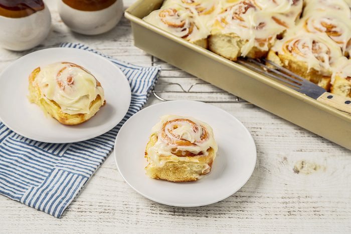A white table displays a large baking dish filled with frosted cinnamon rolls. Two rolls are served on separate white plates, placed on a blue striped cloth. Jars are visible in the background.