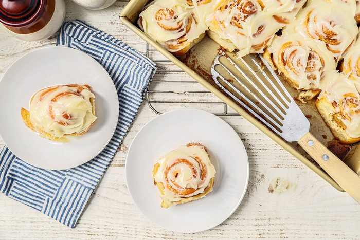 Cinnamon rolls with icing on a white wooden table. Two rolls are on separate plates, beside a pan of more rolls. A striped cloth and a metal spatula are nearby.