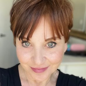 Woman smiling, looking directly at the camera, indoors with blurred background, wearing a black top.